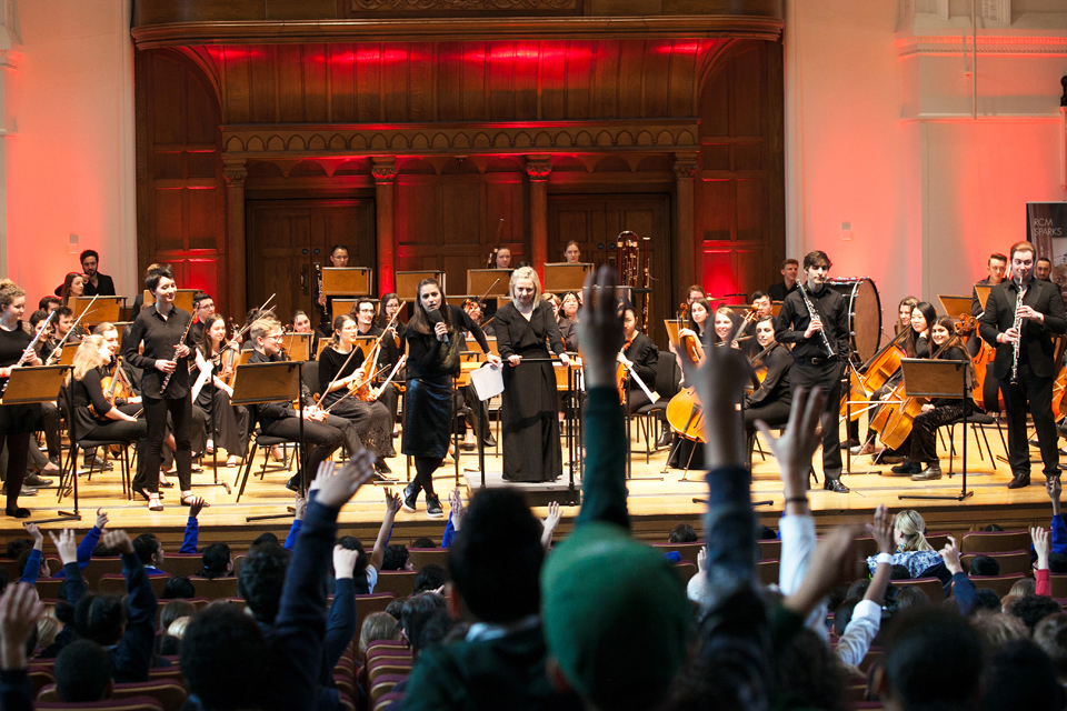 A group of performers, with two women in the centre of an orchestra wearing smart clothes, with a young students putting their hands up in the audience 